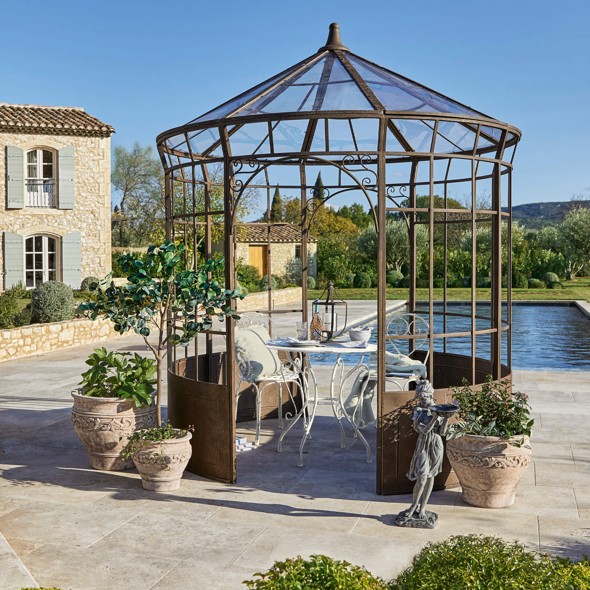 Jardin avec une gloriette en métal rouillé, table et chaises blanches, entourée de pots de plantes