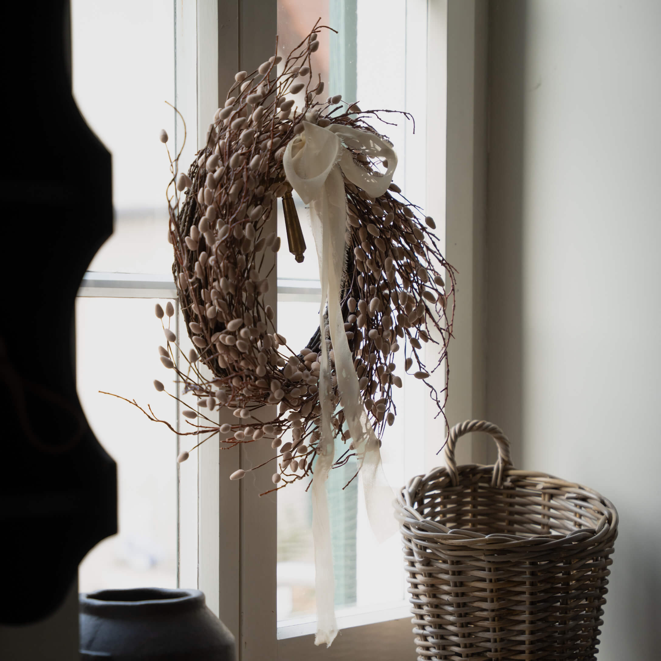 Decoratieve beige krans van takken en knoppen met wit lint aan het raam, rieten mand ernaast