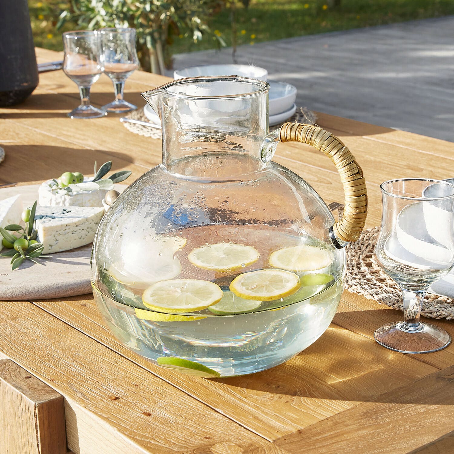 Pichet Stormea Carafe en verre avec anse en rotin sur table en bois, tranches de citron et lime dans l'eau