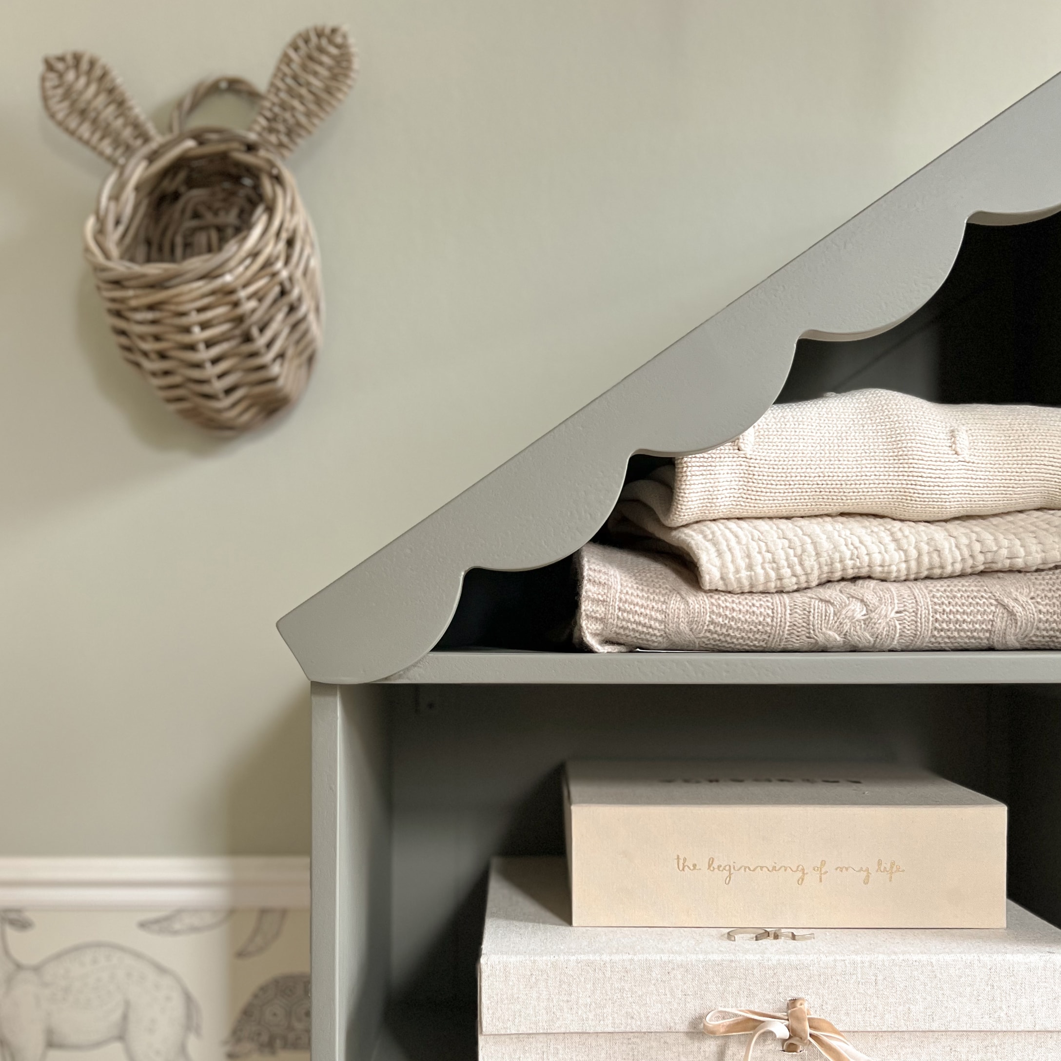 Grijze houten plank met golvend dak, beige truien en decoratieve opbergdozen