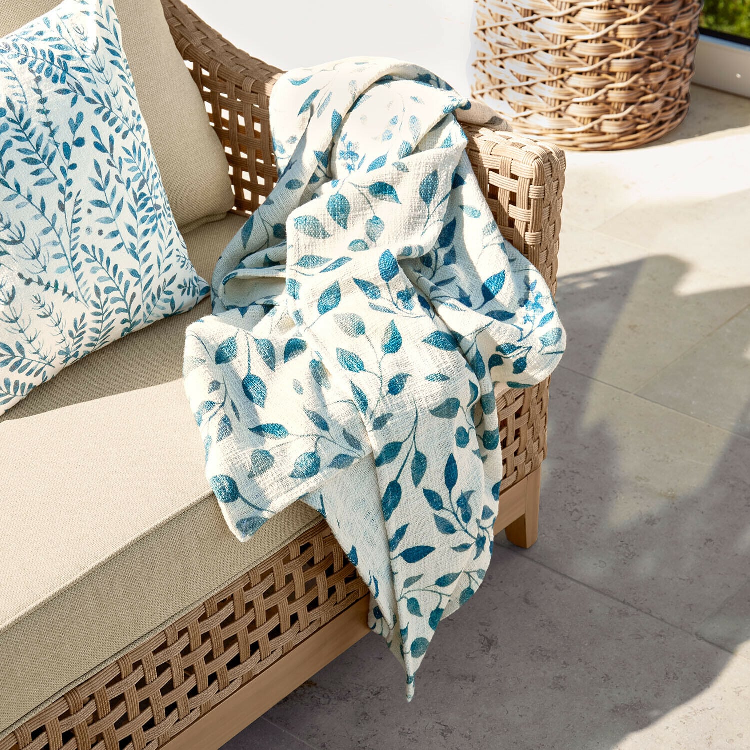 Plaid Ocevara Beige bank met gevlochten rugleuning en crème deken met blauw bladmotief op terras
