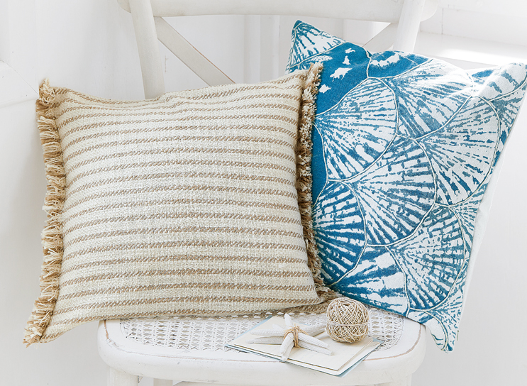 Deux coussins décoratifs sur une chaise blanche, un beige rayé avec franges, l'autre bleu à motif coquillage