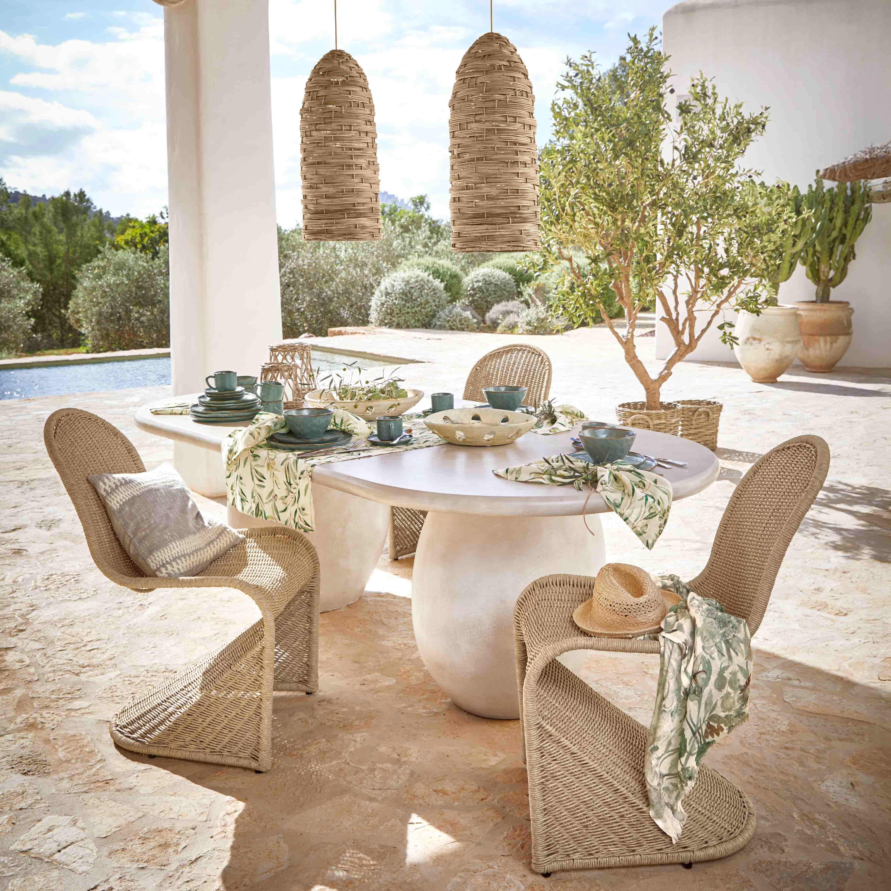Table ovale en pierre blanche avec chaises en osier et vaisselle verte en extérieur près de la piscine