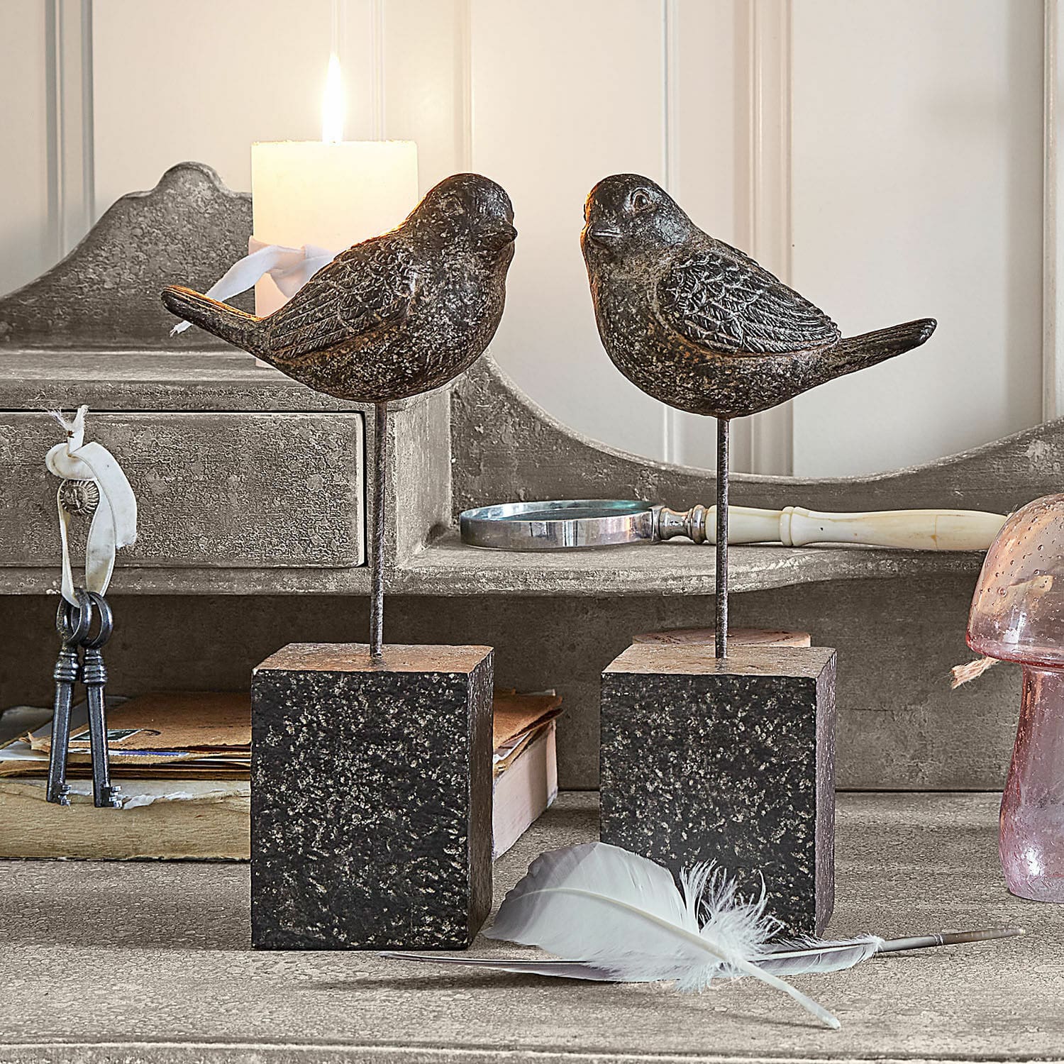 Lot de 2 oiseaux décoratifs Juxivel Oiseaux décoratifs en métal foncé sur socles carrés posés sur une table rustique avec une plume
