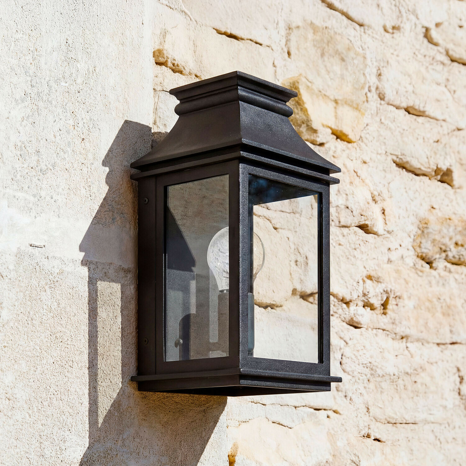 Buitenwandlamp Zélivarne Zwarte metalen wandlamp met helder glas aan lichte stenen muur, klassieke buitenverlichting