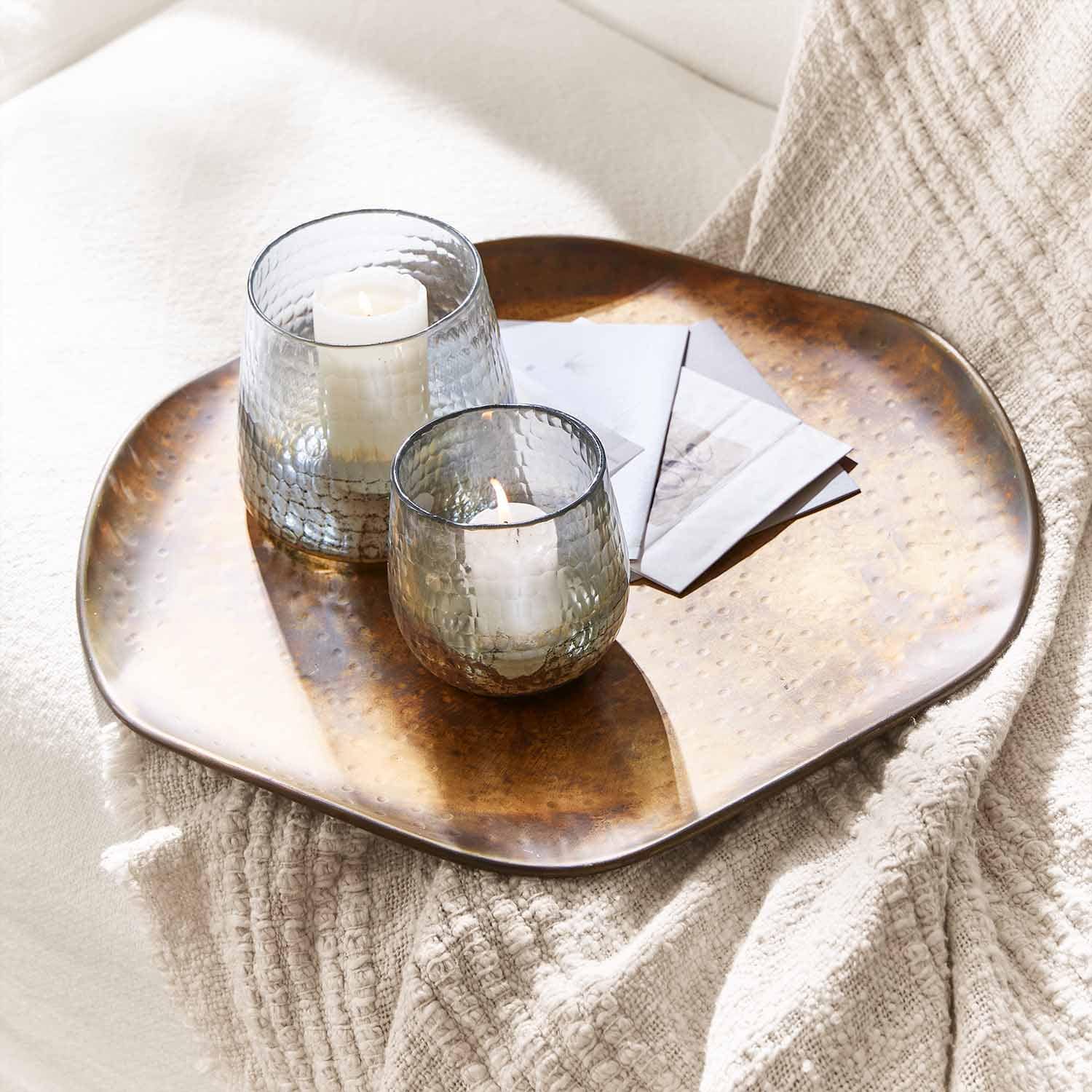 Deux photophores en verre avec bougies blanches sur un plateau en cuivre posé sur une couverture claire