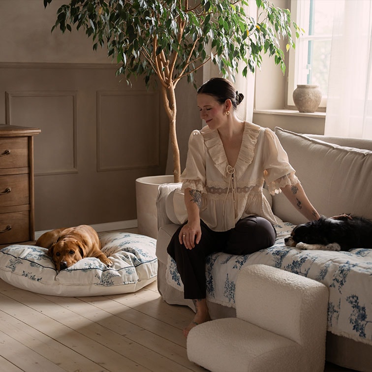 Femme en blouse beige assise sur un canapé avec deux chiens dans un salon chaleureux et lumineux