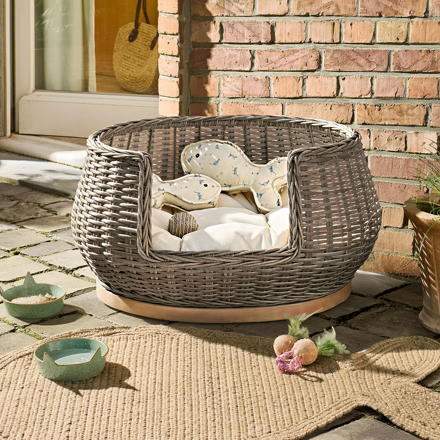 Lit d’intérieur et d’extérieur pour animaux Brinelza Panier rond en osier gris avec coussin crème et jouets pour chien sur terrasse en pierre