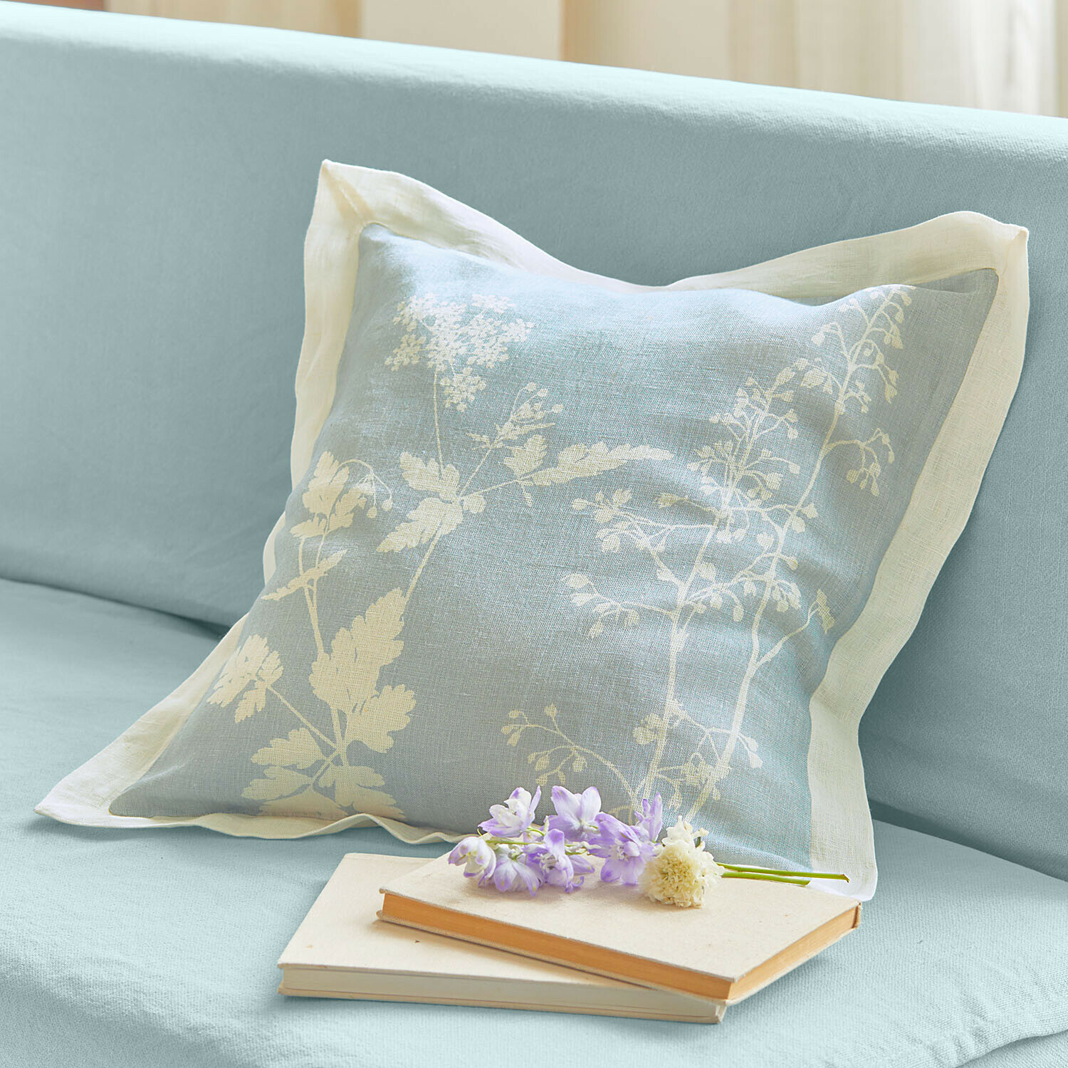 Housse de coussin Hazelwick Coussin en lin bleu clair avec motif floral blanc sur canapé, livres et fleurs posés à côté