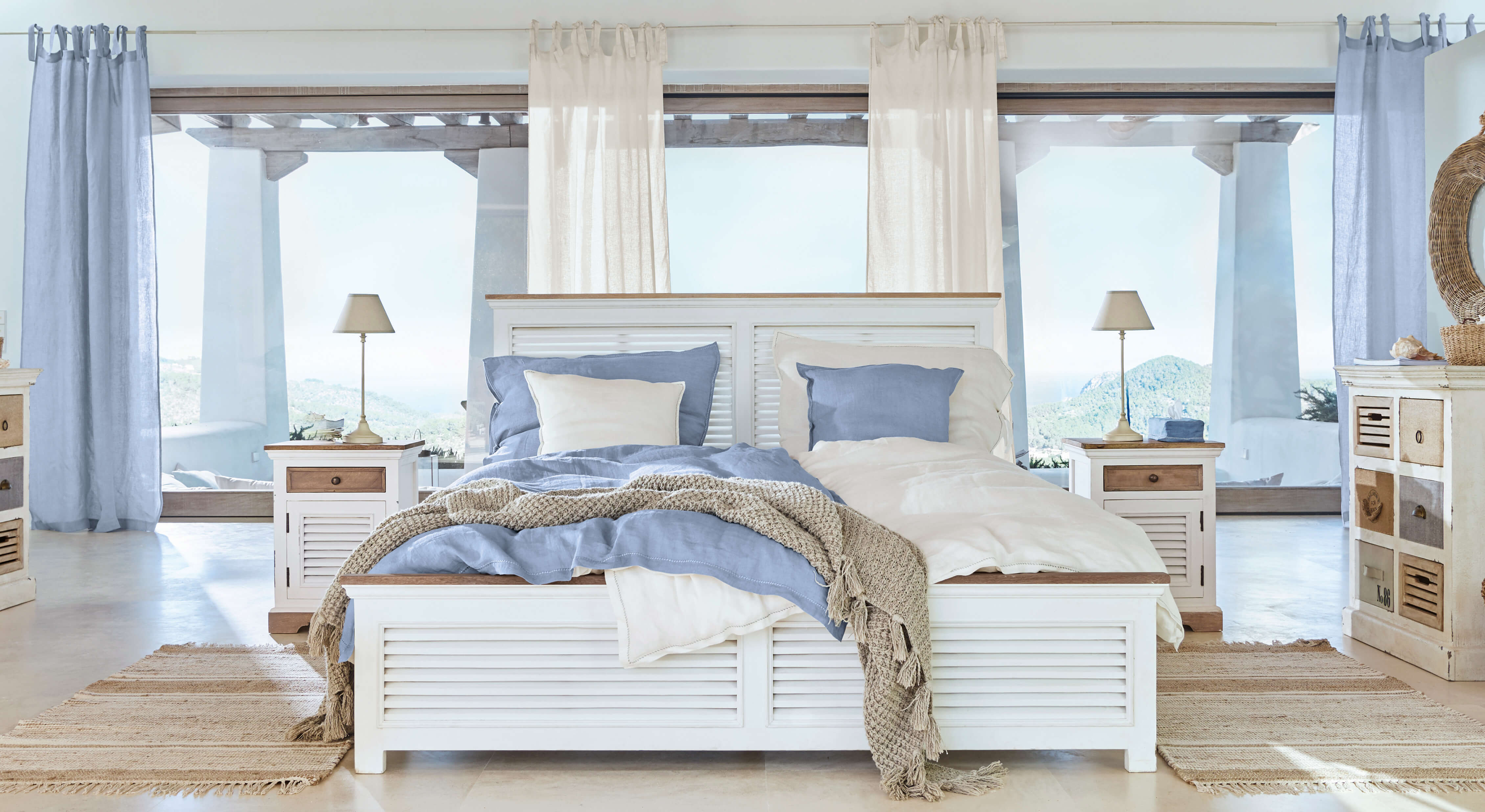 Wit houten bed met blauw-beige beddengoed en natuurlijke vloerkleed in lichte slaapkamer met uitzicht