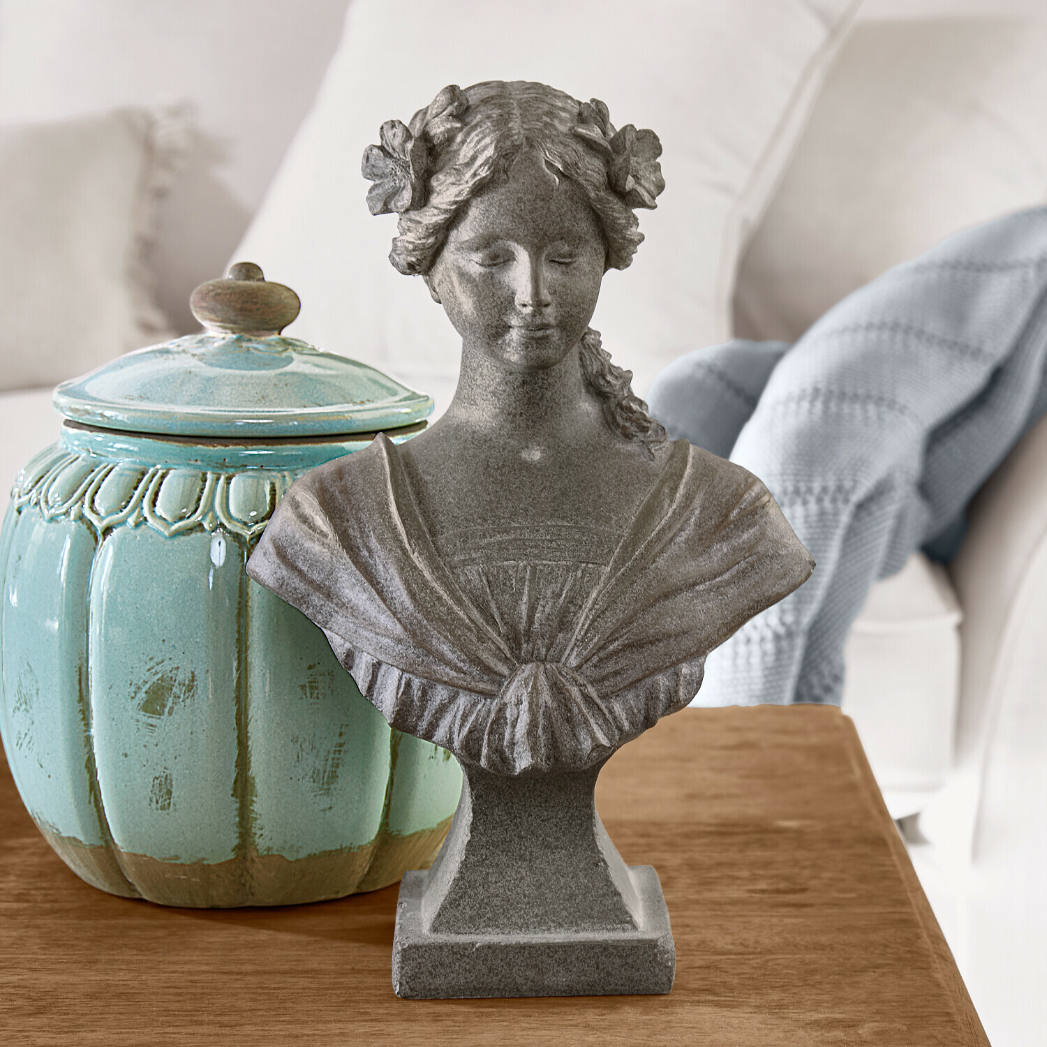 Buste féminin en pierre grise avec fleurs dans les cheveux sur table en bois à côté d’un pot en céramique turquoise