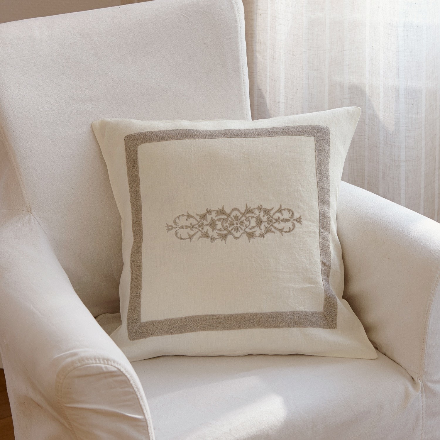 Coussin décoratif en lin beige avec motif brodé et bordure sur fauteuil crème