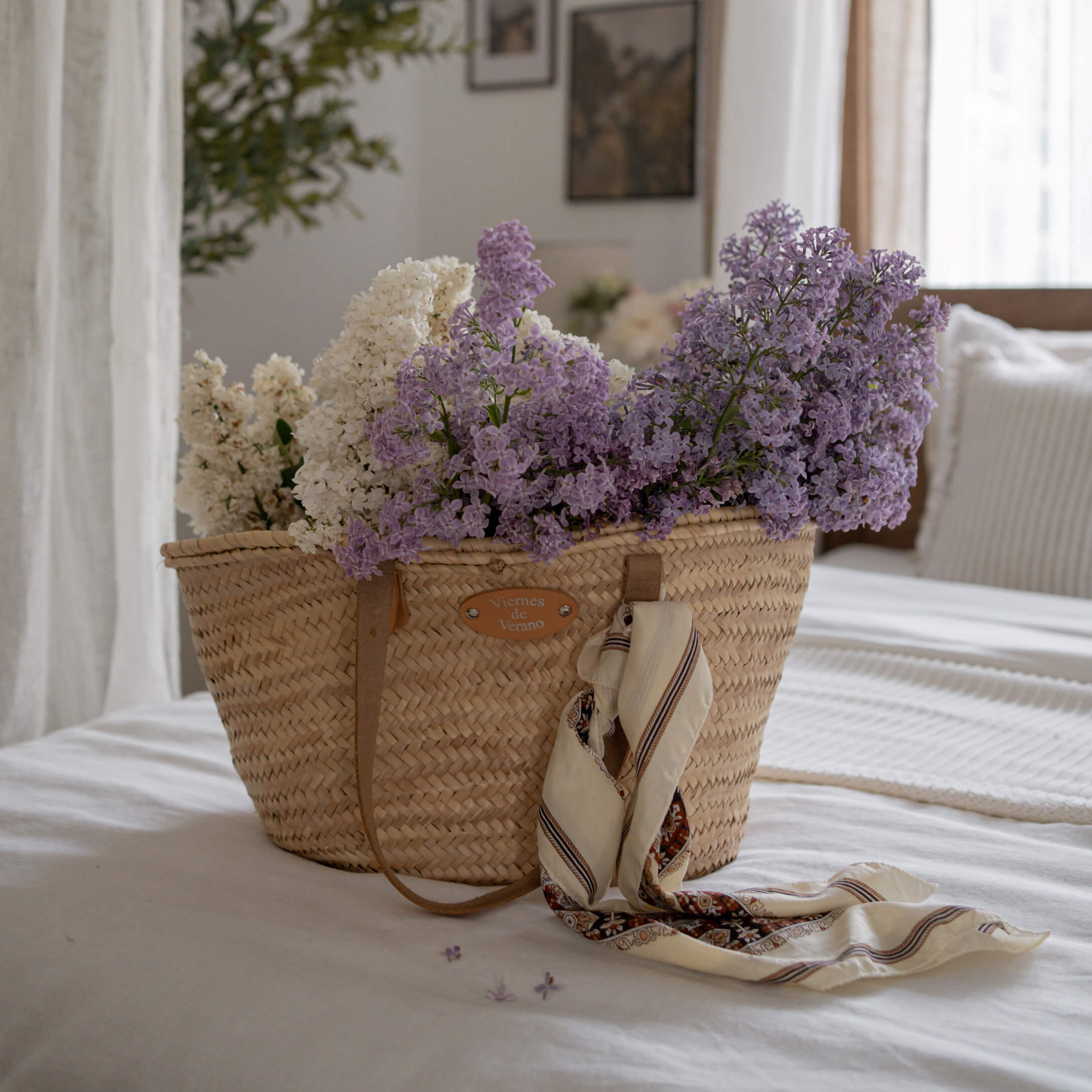 Panier en osier avec fleurs blanches et violettes et foulard posé sur un lit blanc dans une chambre lumineuse
