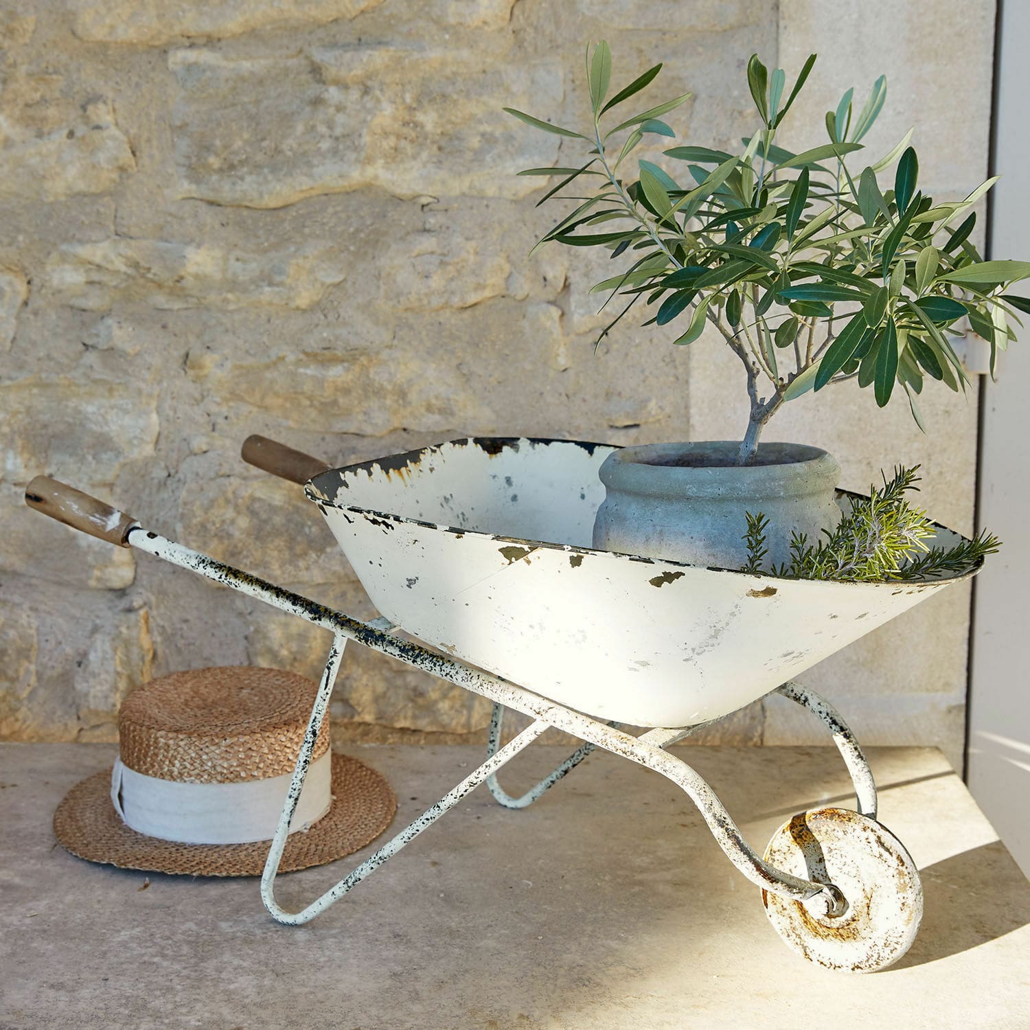 Brouette décorative Padoux Brouette en métal blanc vieilli avec pot et plantes vertes devant un mur en pierre
