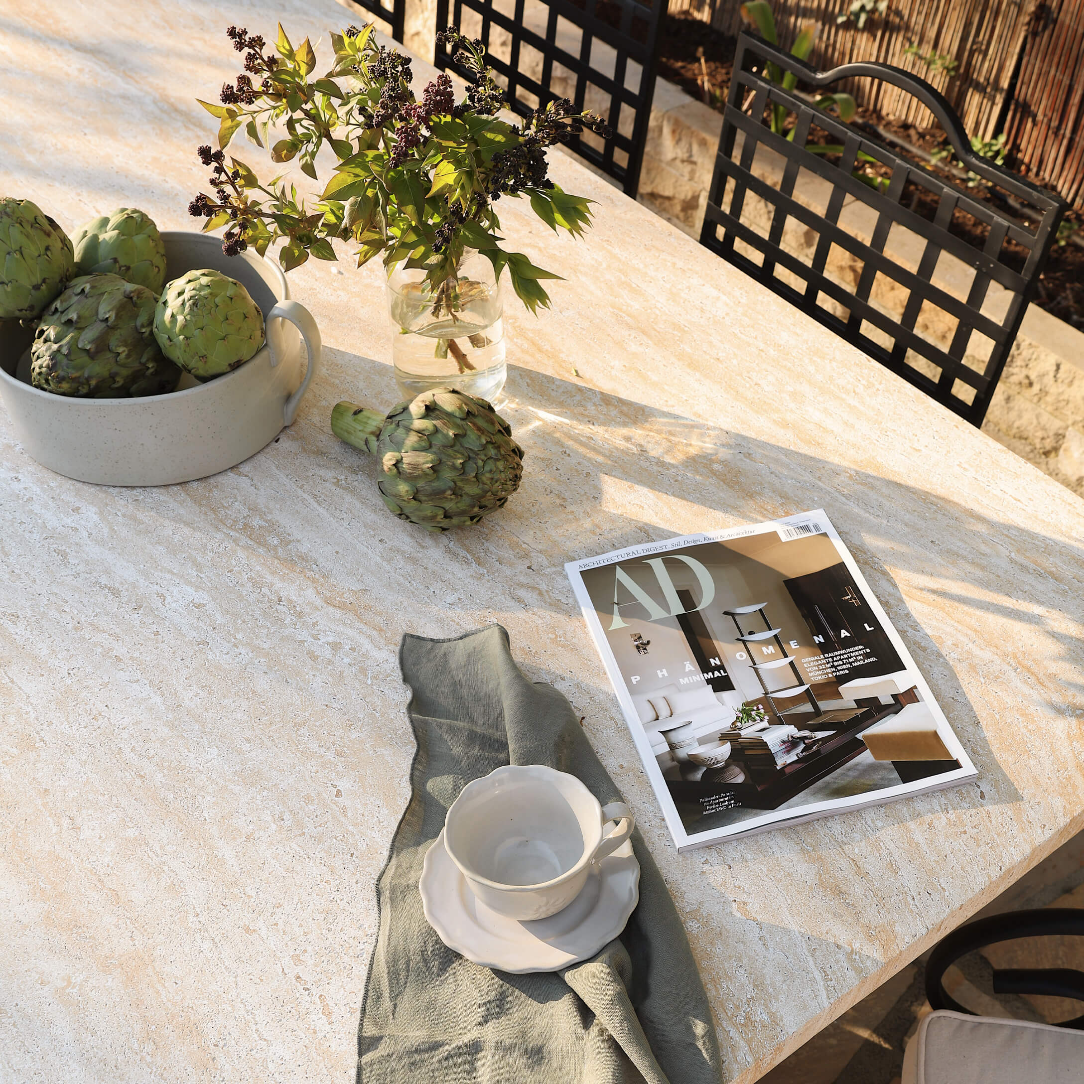 Lichte stenen tafel met groene artisjokken, witte kop en woonmagazine buiten