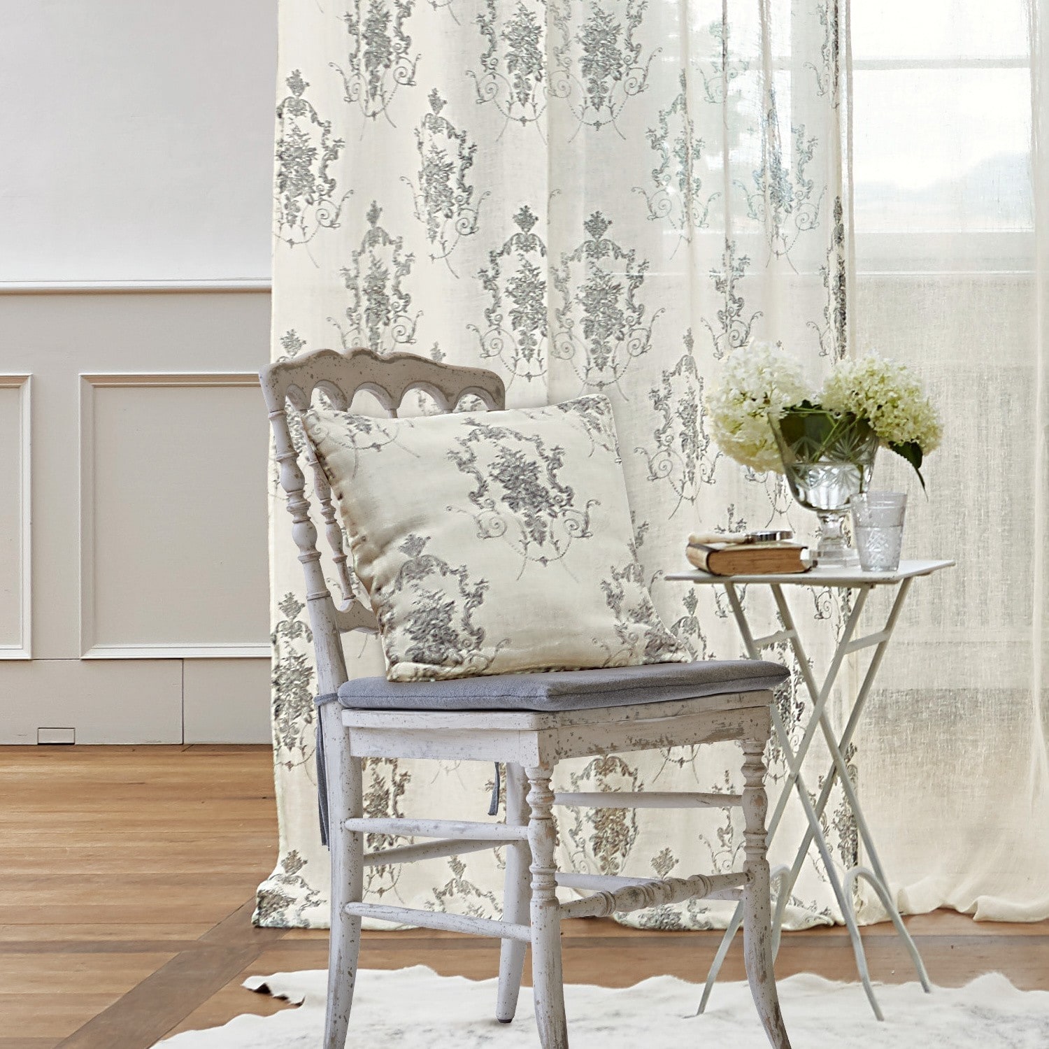Kussenhoes Dinan Witte houten stoel met grijs kussen en decoratief kussen met bloemenpatroon, bijzettafel met bloemen