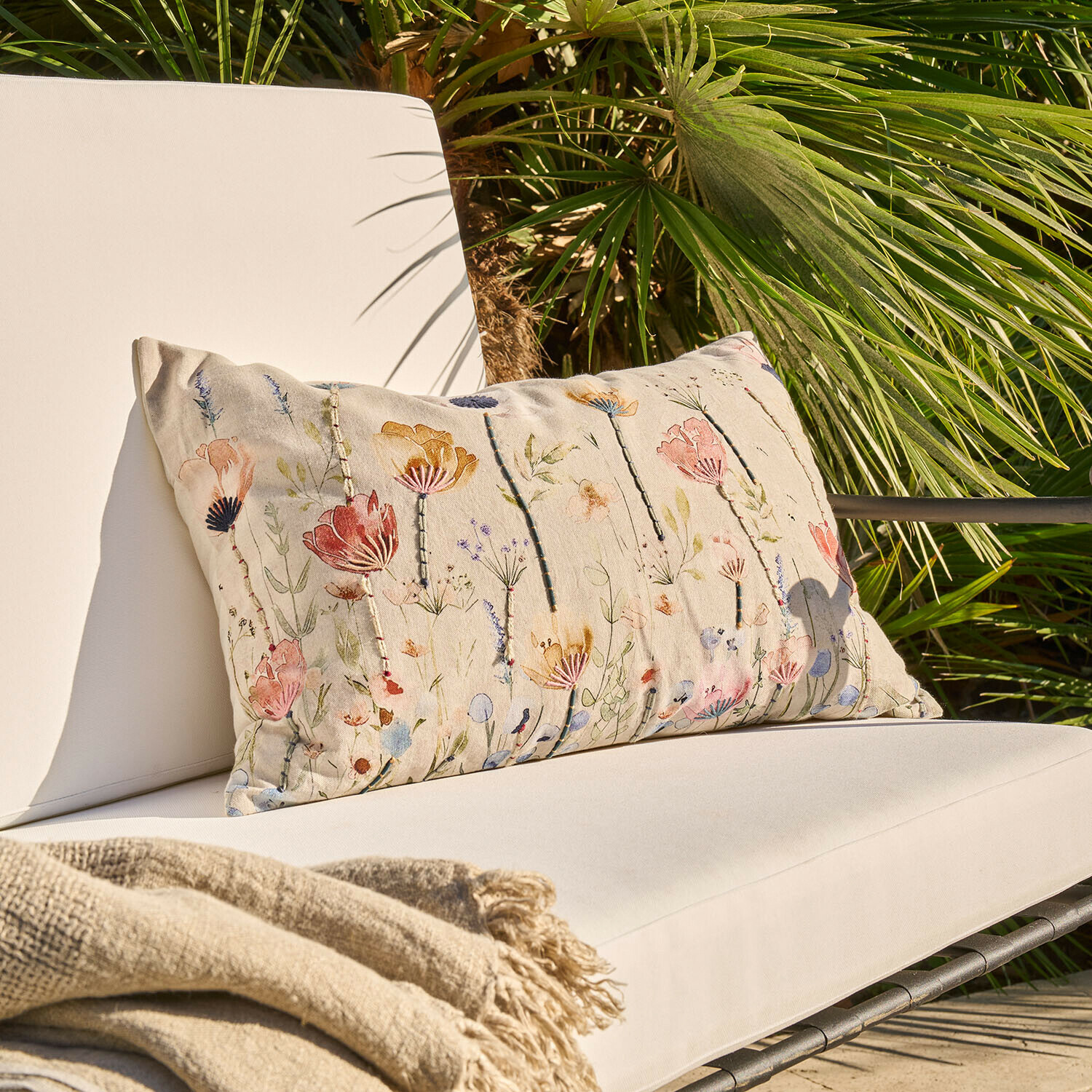 Beige sierkussen met bloemenpatroon op lichte tuinstoel met beige deken