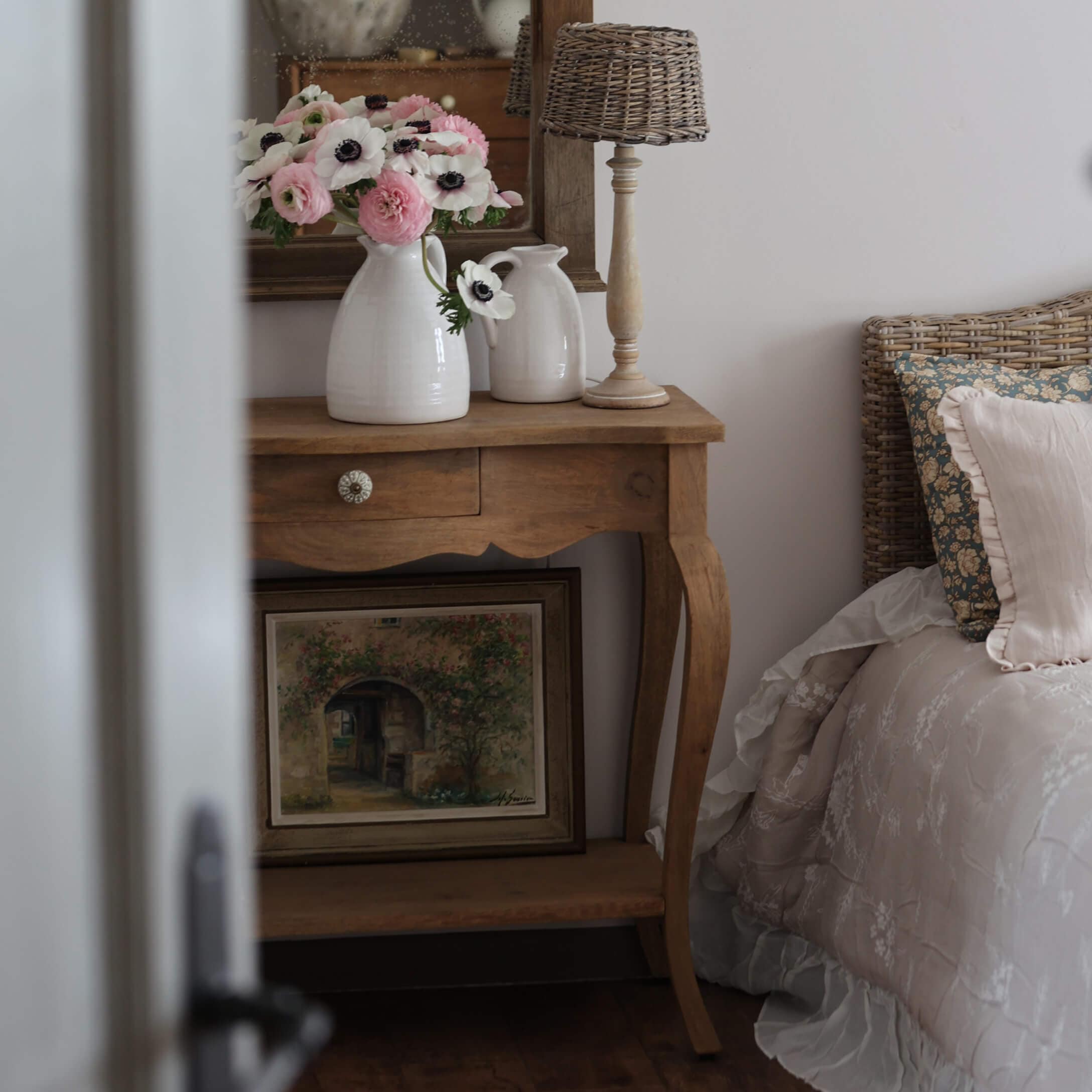 Houten tafeltje met witte keramische kannen en bloemen naast bed met gevlochten hoofdeinde