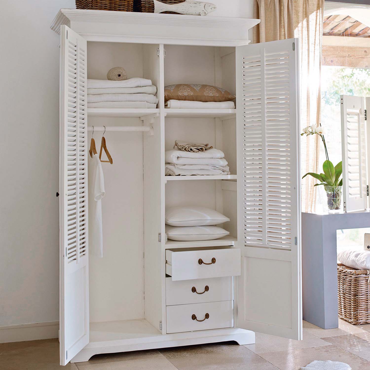 Armoire en bois blanc ouverte avec textiles blancs pliés et coussins dans une pièce lumineuse
