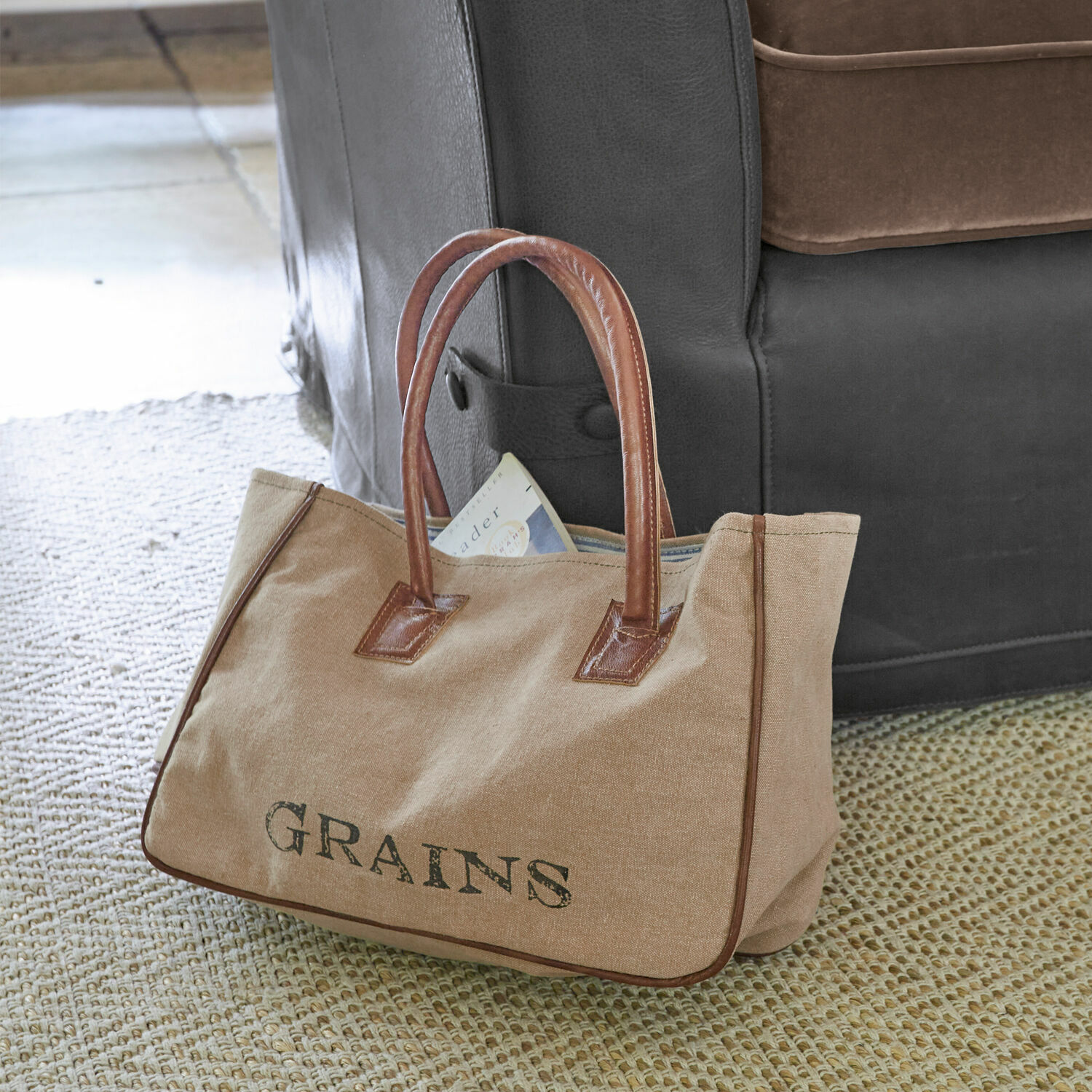 Tas Grains Beige stoffen tas met bruine leren handvatten en tekst GRAINS op tapijt naast bank