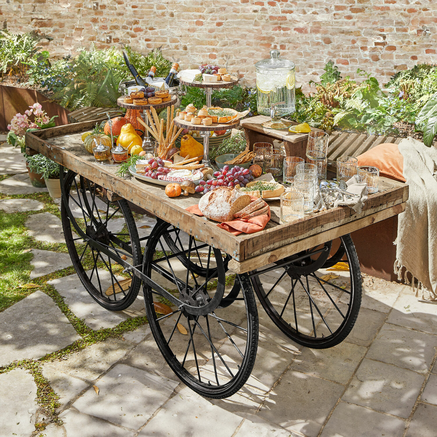 Desserte Annabel Table de jardin en bois et métal avec roues, garnie de snacks et boissons en extérieur