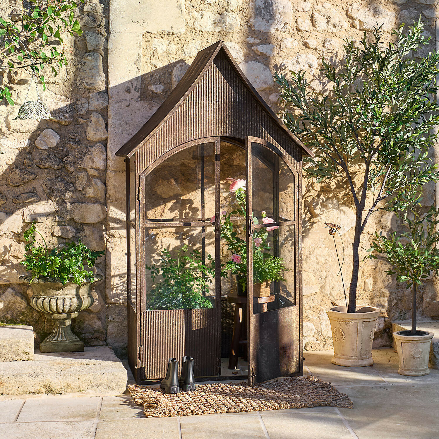 Serre sur pieds Parvignon Serre de jardin en métal brun style vintage avec plantes et pots devant un mur en pierre ensoleillé