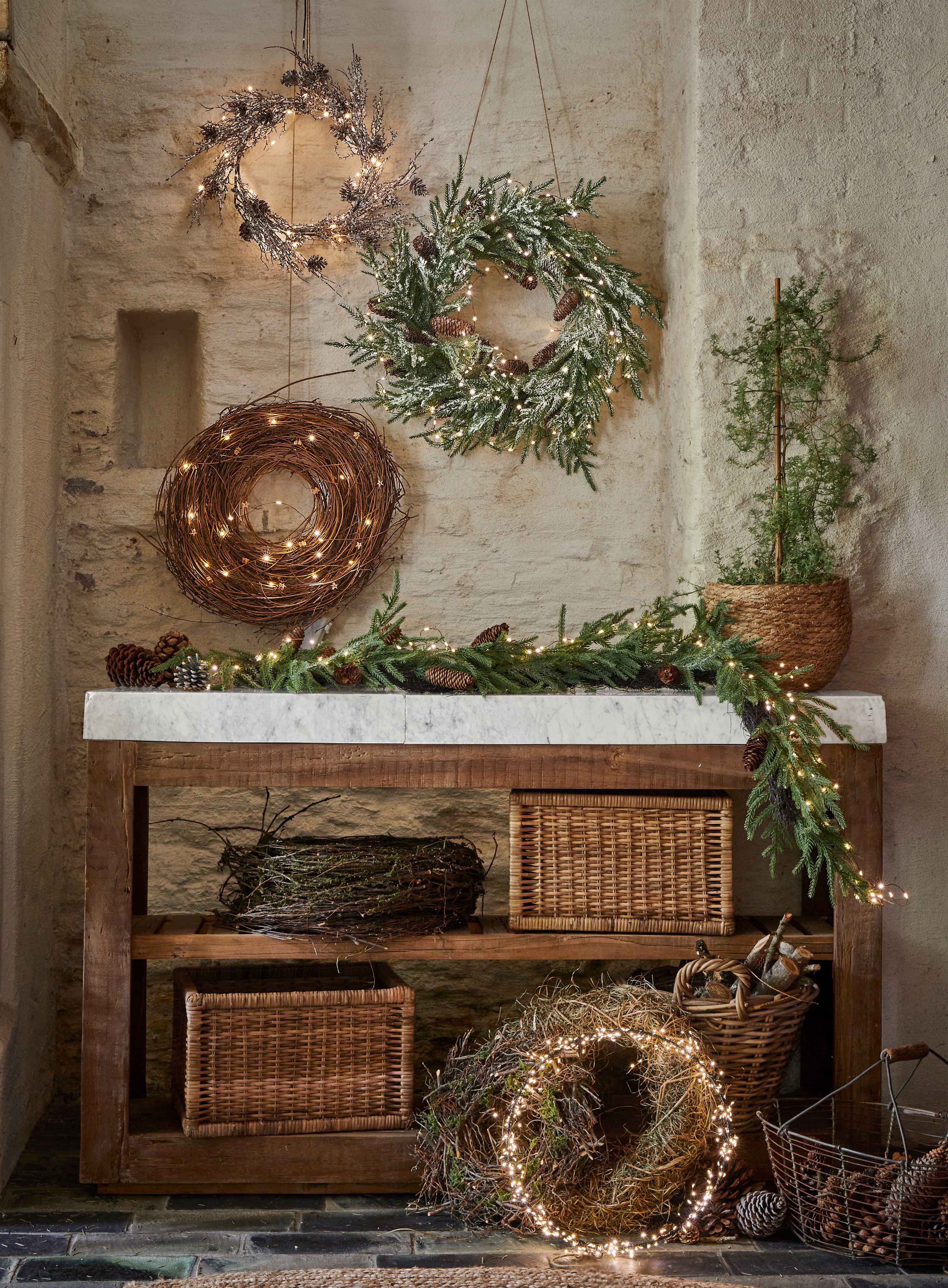 Couronnes décoratives de Noël en branches, pommes de pin et lumières sur une table en bois dans un intérieur rustique