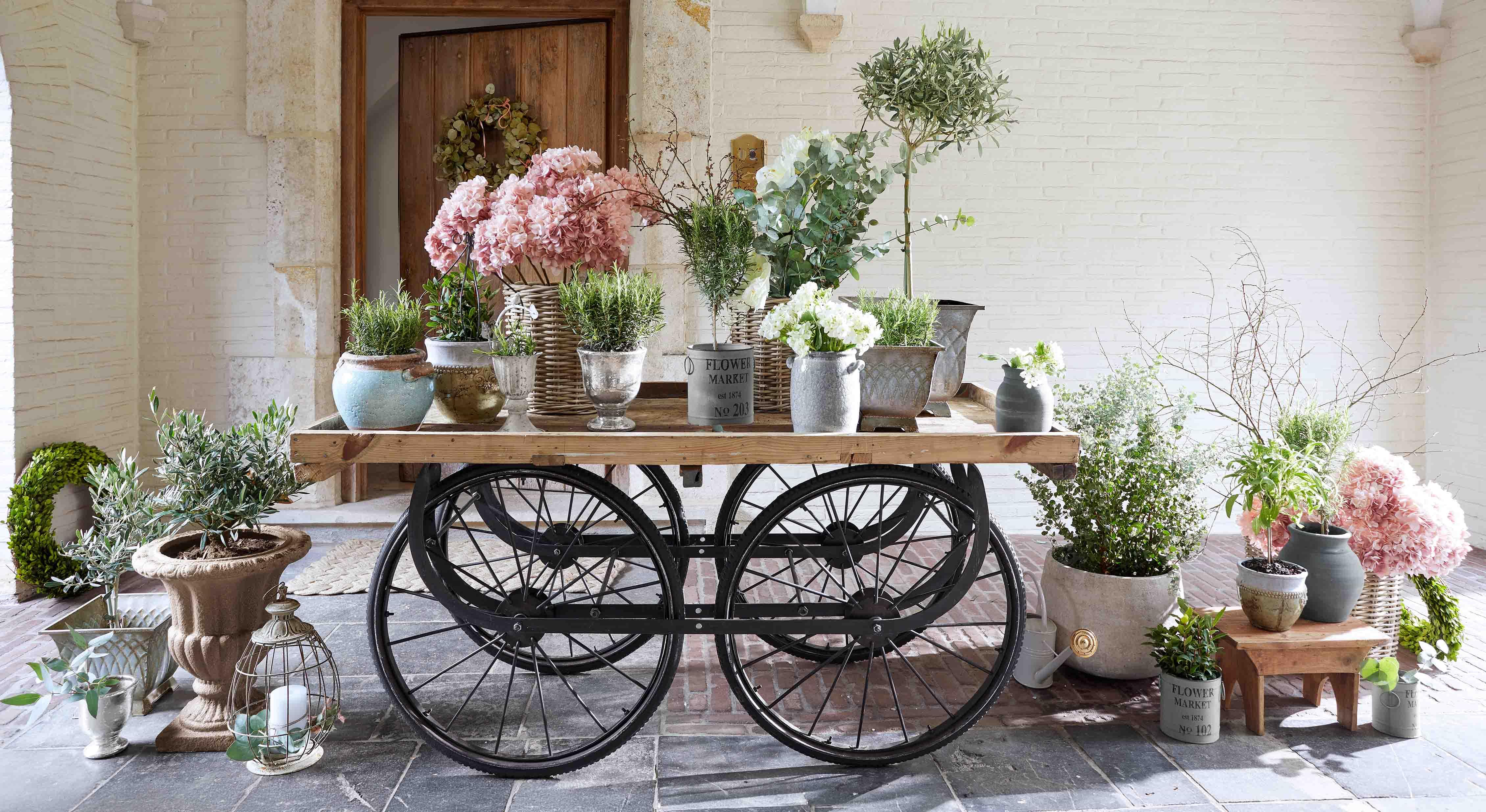 Houten kar met zwarte wielen en diverse bloempotten met roze en witte bloemen