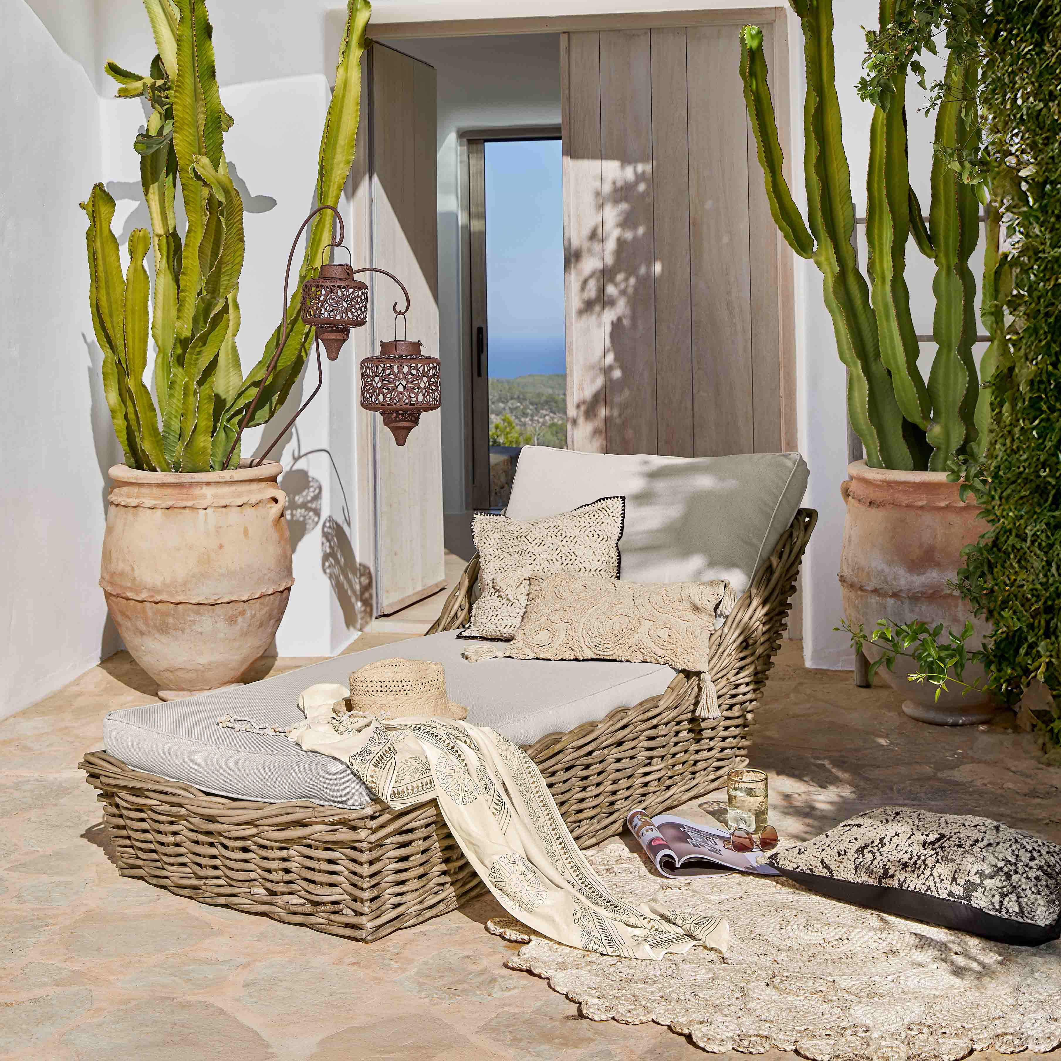 Chaise longue en osier avec coussins gris clair sur terrasse décorée de cactus en pot