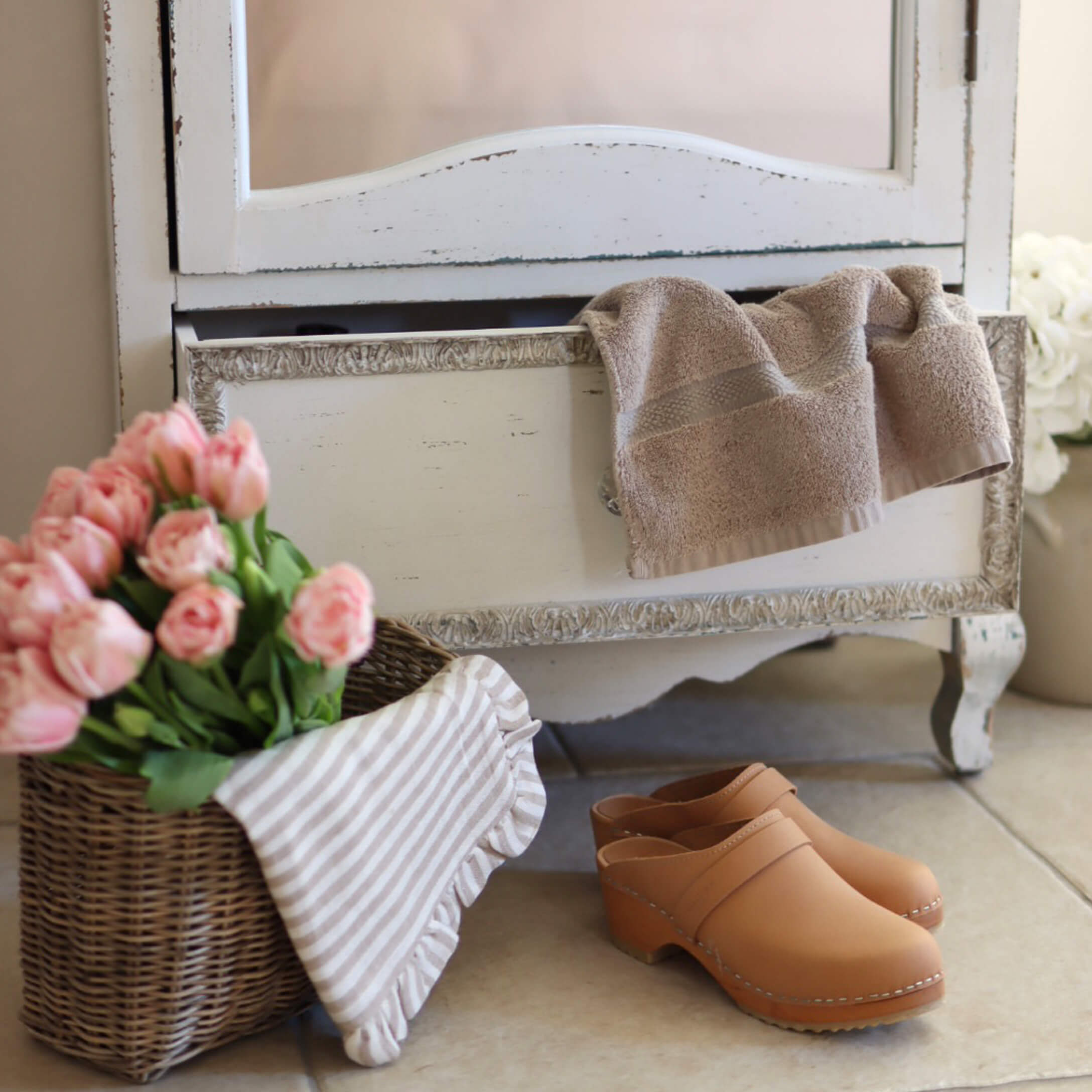 Sabots en cuir beige posés au sol près d'un panier en osier avec des fleurs roses et une serviette