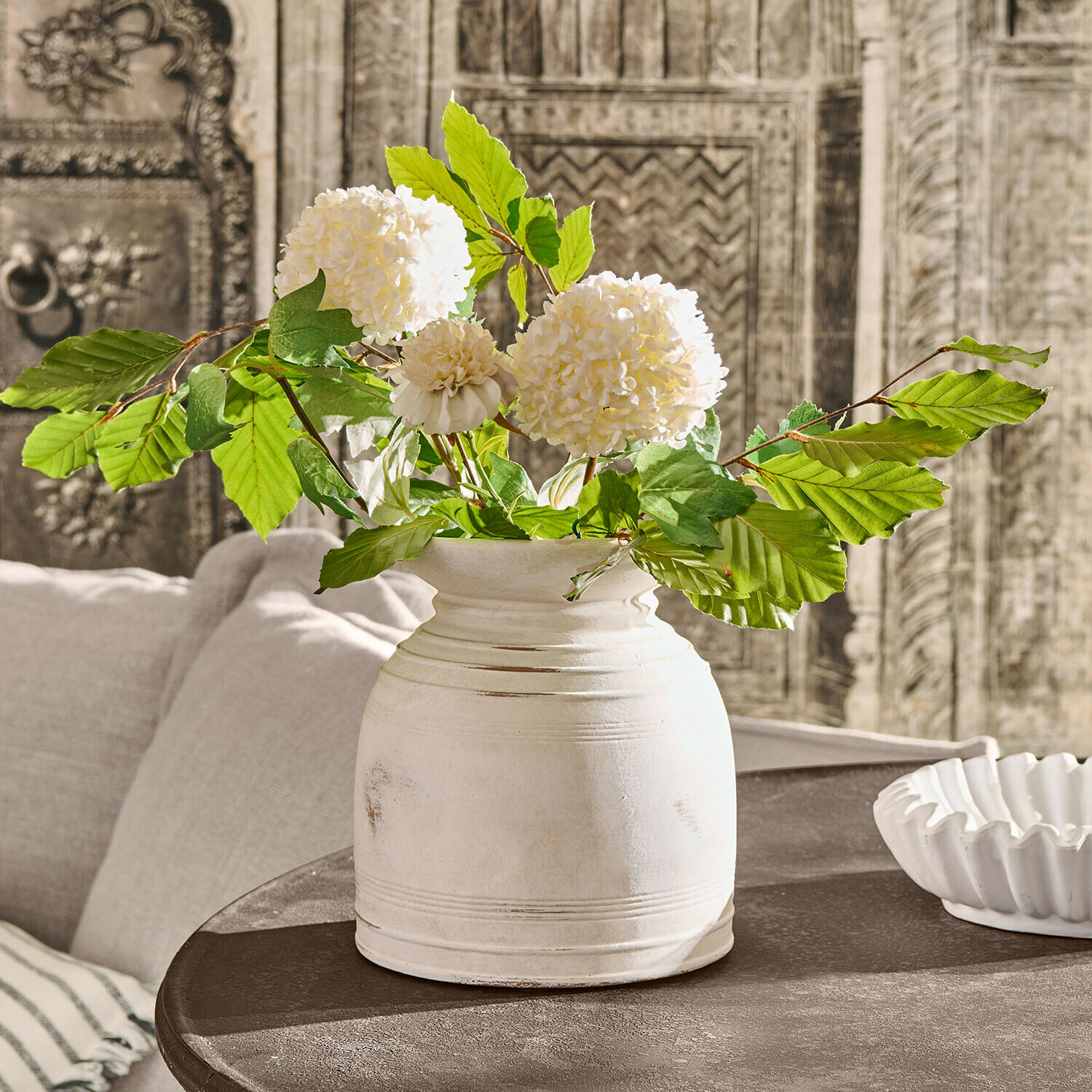 Vase Niverana Vase en céramique blanche avec feuillage vert et fleurs crème sur table sombre style vintage